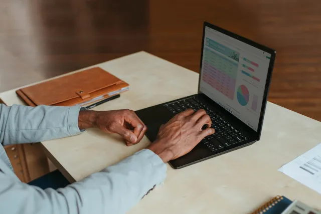 A person types on a laptop showing a spreadsheet and charts, beside a leather folio on a pale desk.