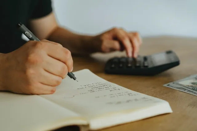 A person writes figures in a notebook beside a calculator and a few dollar bills.