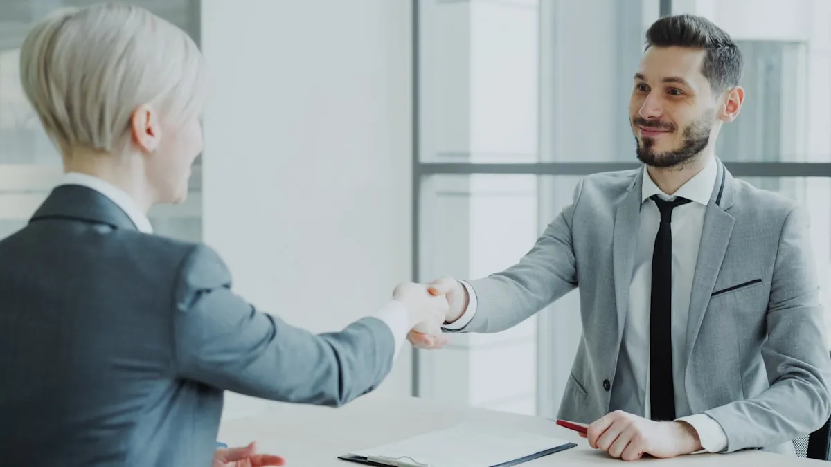 Two people in gray suits shake hands across an office desk.