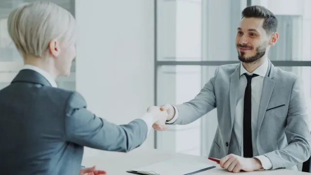 Two people in gray suits shake hands across an office desk.