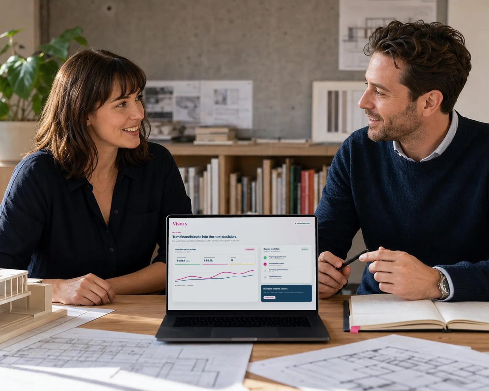 Two architects smile at each other across a studio table with a building model, drawings, and a laptop.