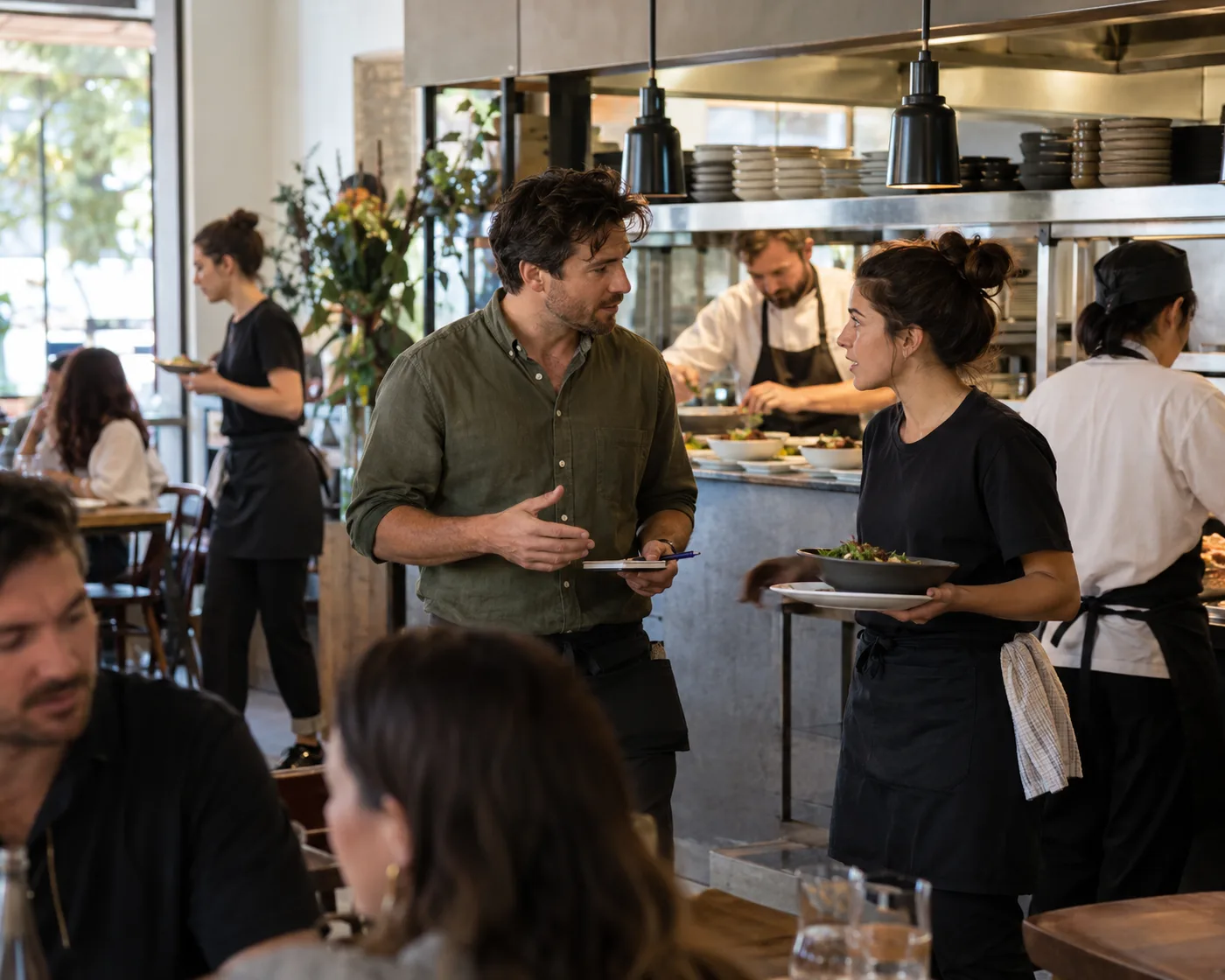 A restaurant manager talks with a server at the kitchen pass during a busy lunch service.