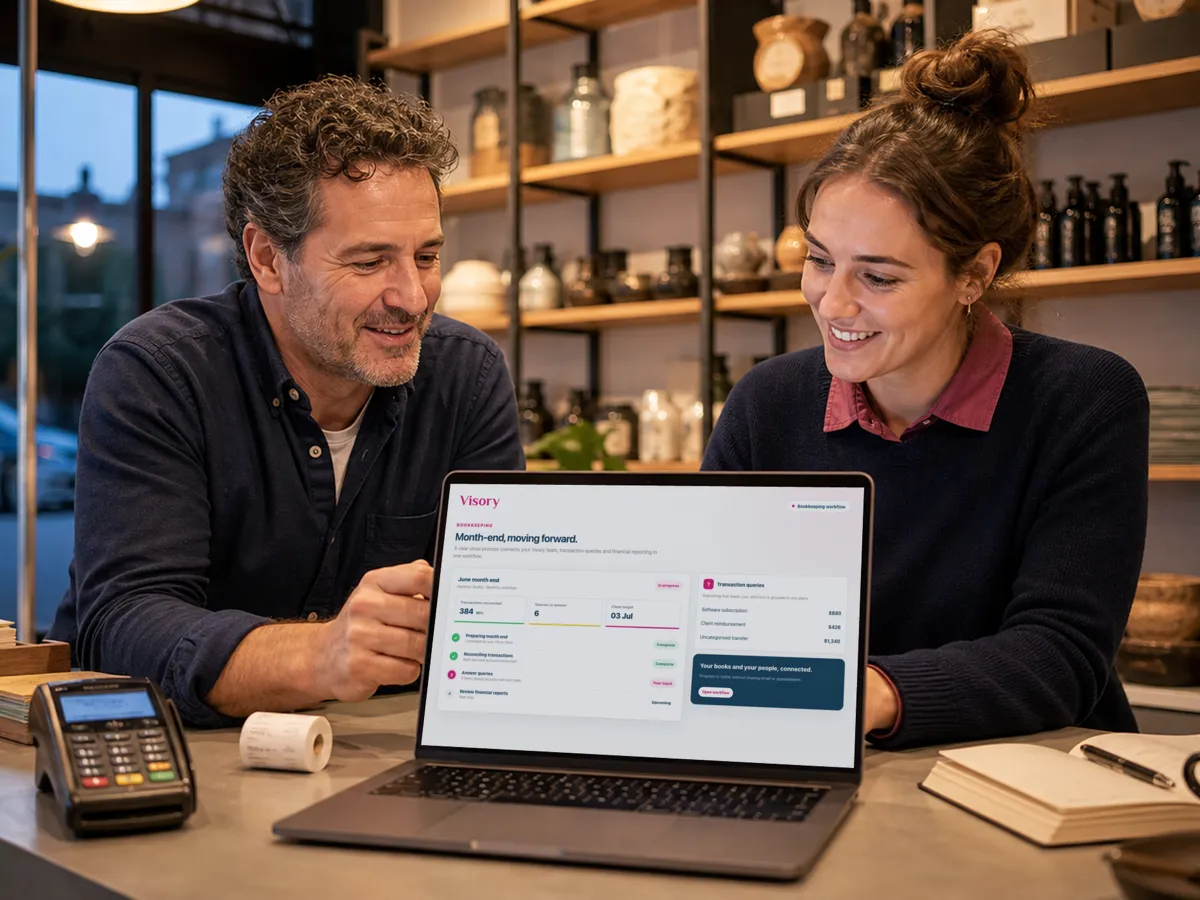 An owner and a colleague review month-end figures on a laptop at the counter, a card terminal beside them.