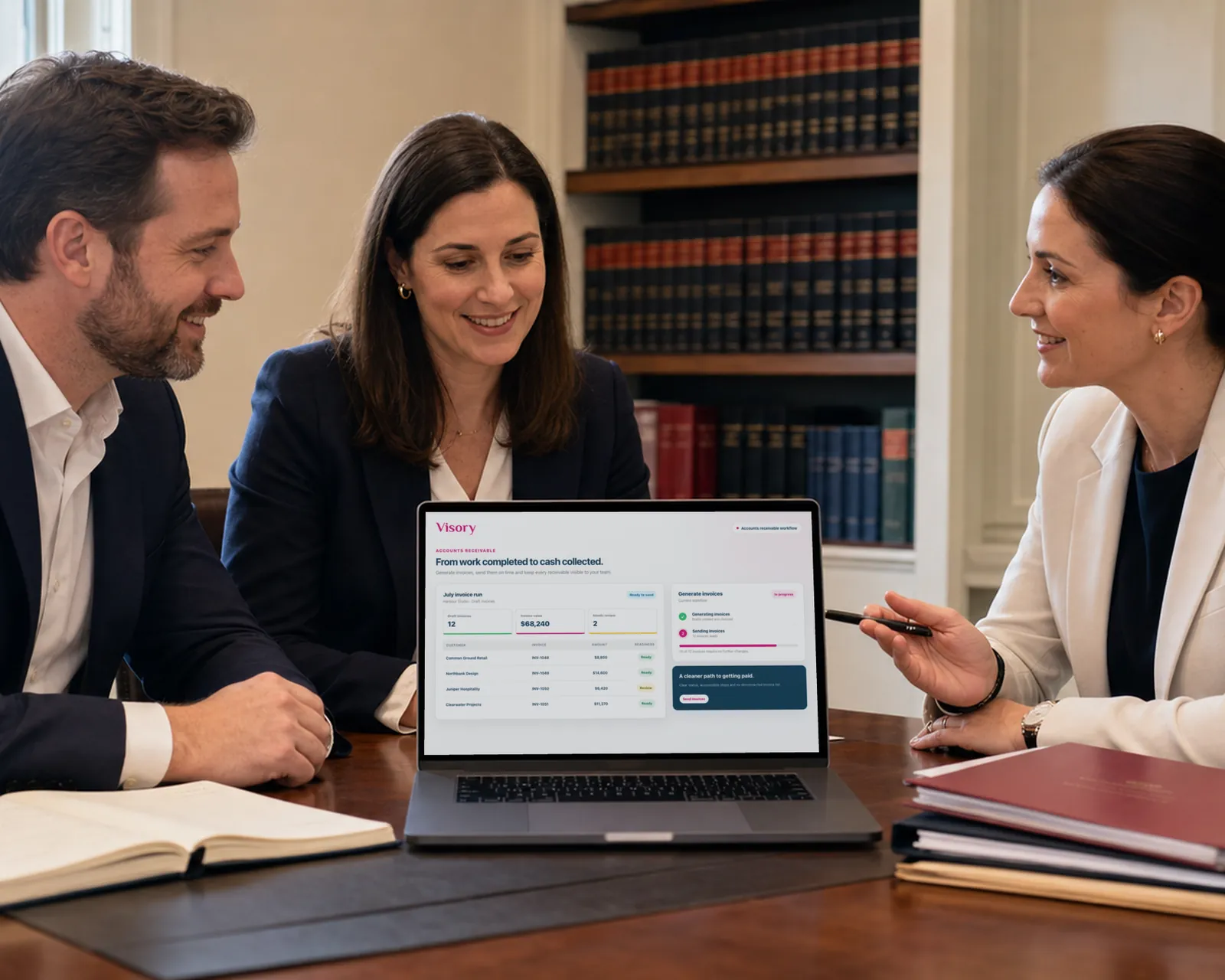 Three attorneys at a meeting table in a book-lined office smile over a laptop showing an invoice run.