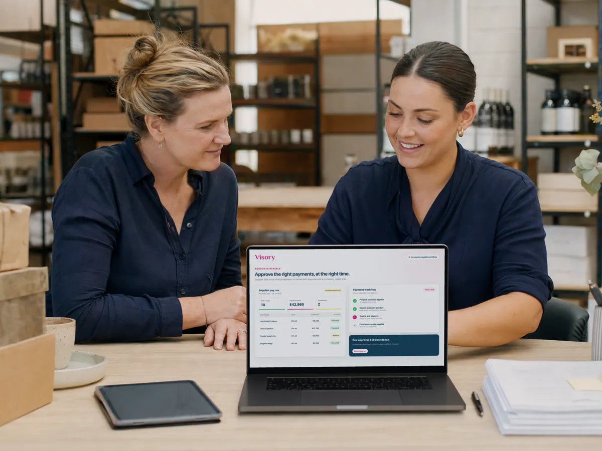 Two colleagues review a supplier pay run on a laptop in a stockroom lined with shelves and parcels.