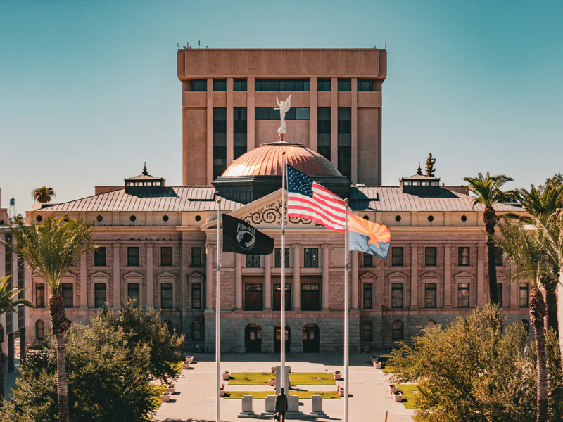 The Arizona State Capitol in Phoenix, its copper dome topped by a winged figure, with flags flying over the palm-lined plaza in front.