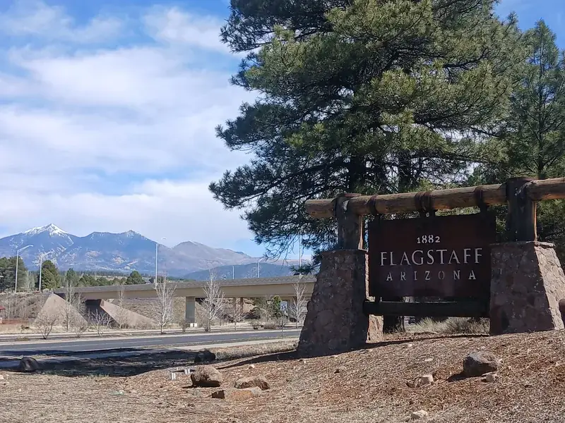 The Flagstaff welcome sign under ponderosa pines, with the snow-capped San Francisco Peaks behind.