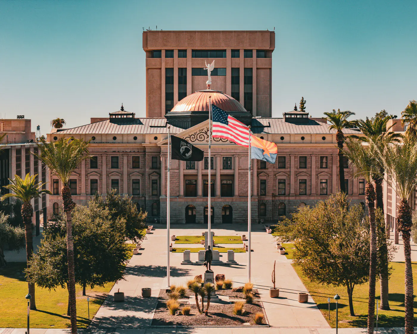 The Arizona State Capitol in Phoenix, its copper dome topped by a winged figure, with flags flying over the palm-lined plaza in front.