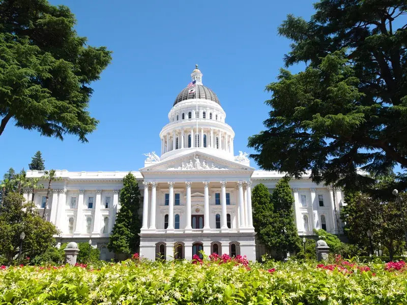 The California State Capitol in Sacramento, its white dome above flowering gardens and cedars under a clear blue sky.