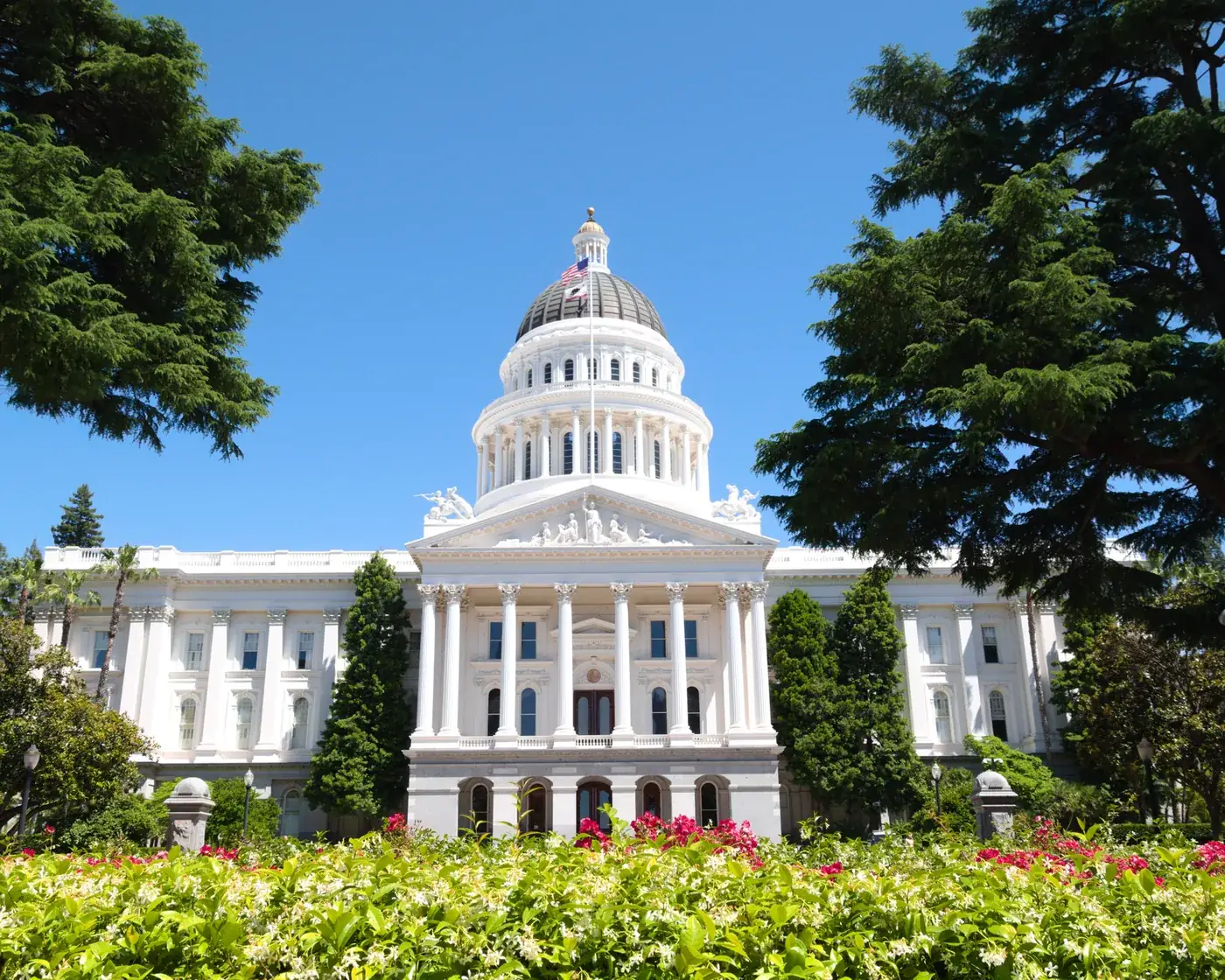 The California State Capitol in Sacramento, its white dome above flowering gardens and cedars under a clear blue sky.