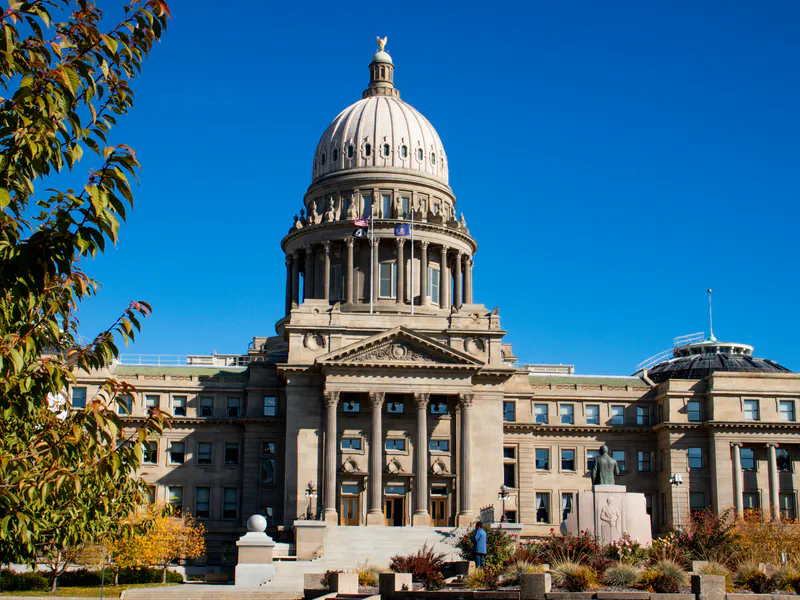The Idaho State Capitol in Boise, its sandstone dome and columned front under a deep blue sky, framed by autumn leaves.