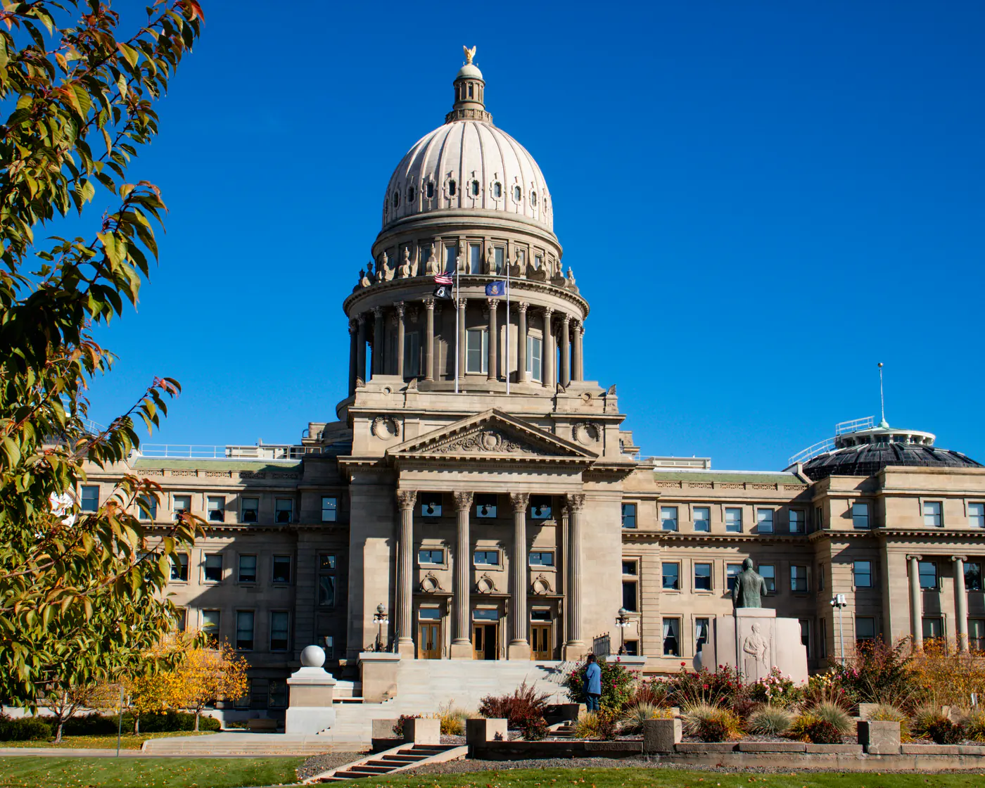 The Idaho State Capitol in Boise, its sandstone dome and columned front under a deep blue sky, framed by autumn leaves.