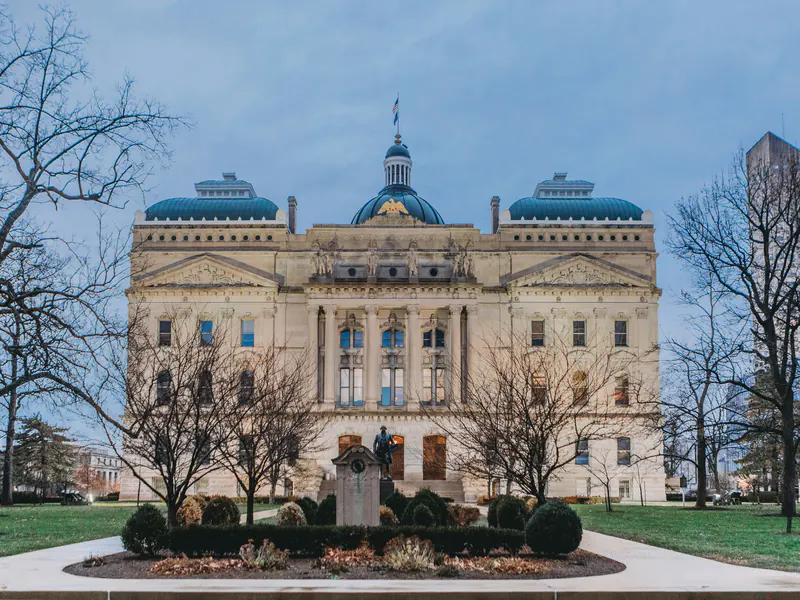 The Indiana Statehouse in Indianapolis at dusk, its central dome and green copper roofs behind bare winter trees and a statue on the lawn.