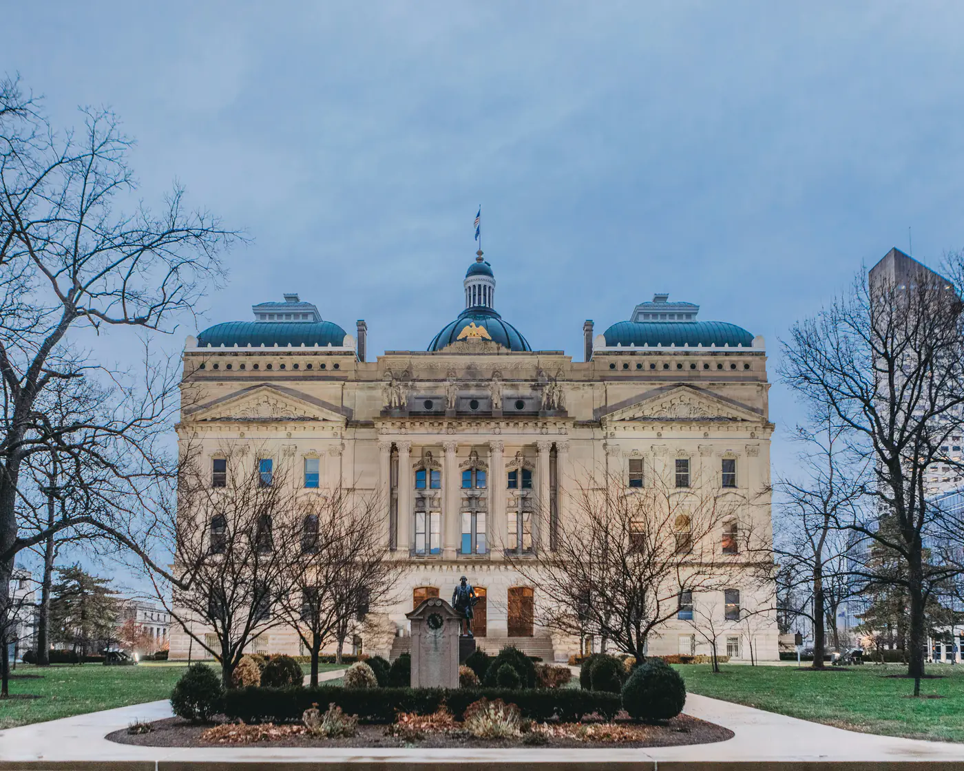 The Indiana Statehouse in Indianapolis at dusk, its central dome and green copper roofs behind bare winter trees and a statue on the lawn.