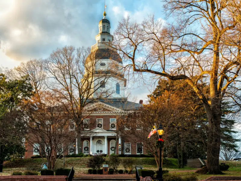 The Maryland State House in Annapolis, its white wooden dome rising above bare trees lit by late sun, with wreaths hung in the windows.