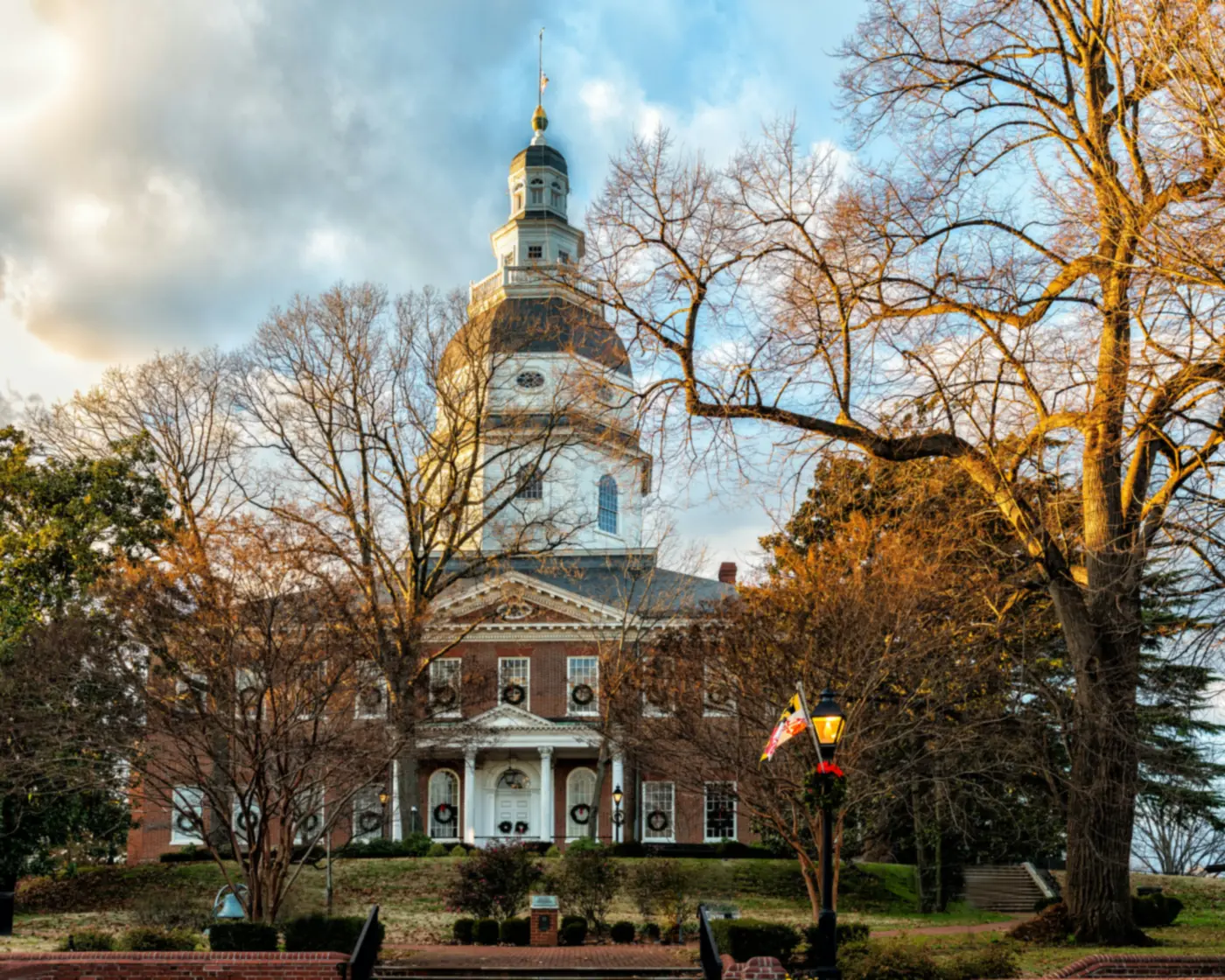 The Maryland State House in Annapolis, its white wooden dome rising above bare trees lit by late sun, with wreaths hung in the windows.