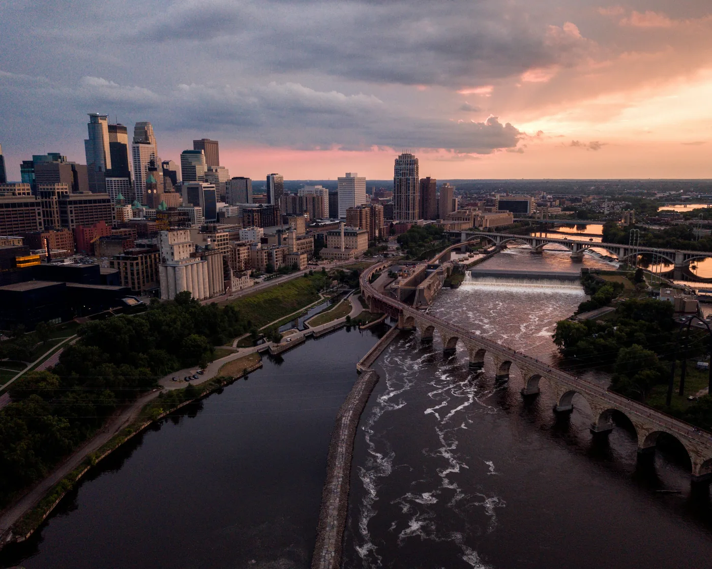 Downtown Minneapolis, the Stone Arch Bridge and St. Anthony Falls on the Mississippi River under a sunset sky.