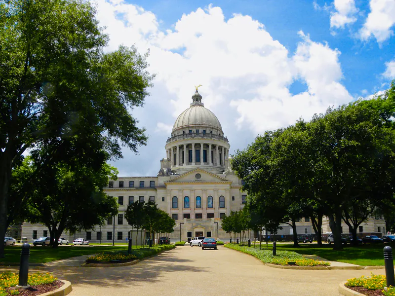 The Mississippi State Capitol in Jackson, its dome topped by a gilded eagle, at the end of a tree-lined drive on a summer day.