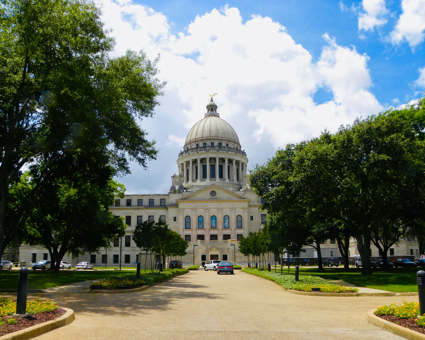 The Mississippi State Capitol in Jackson, its dome topped by a gilded eagle, at the end of a tree-lined drive on a summer day.