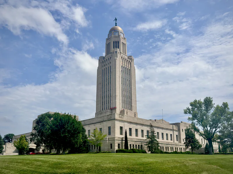 The Nebraska State Capitol in Lincoln, its limestone tower and gold dome rising above a green lawn and trees under scattered cloud.