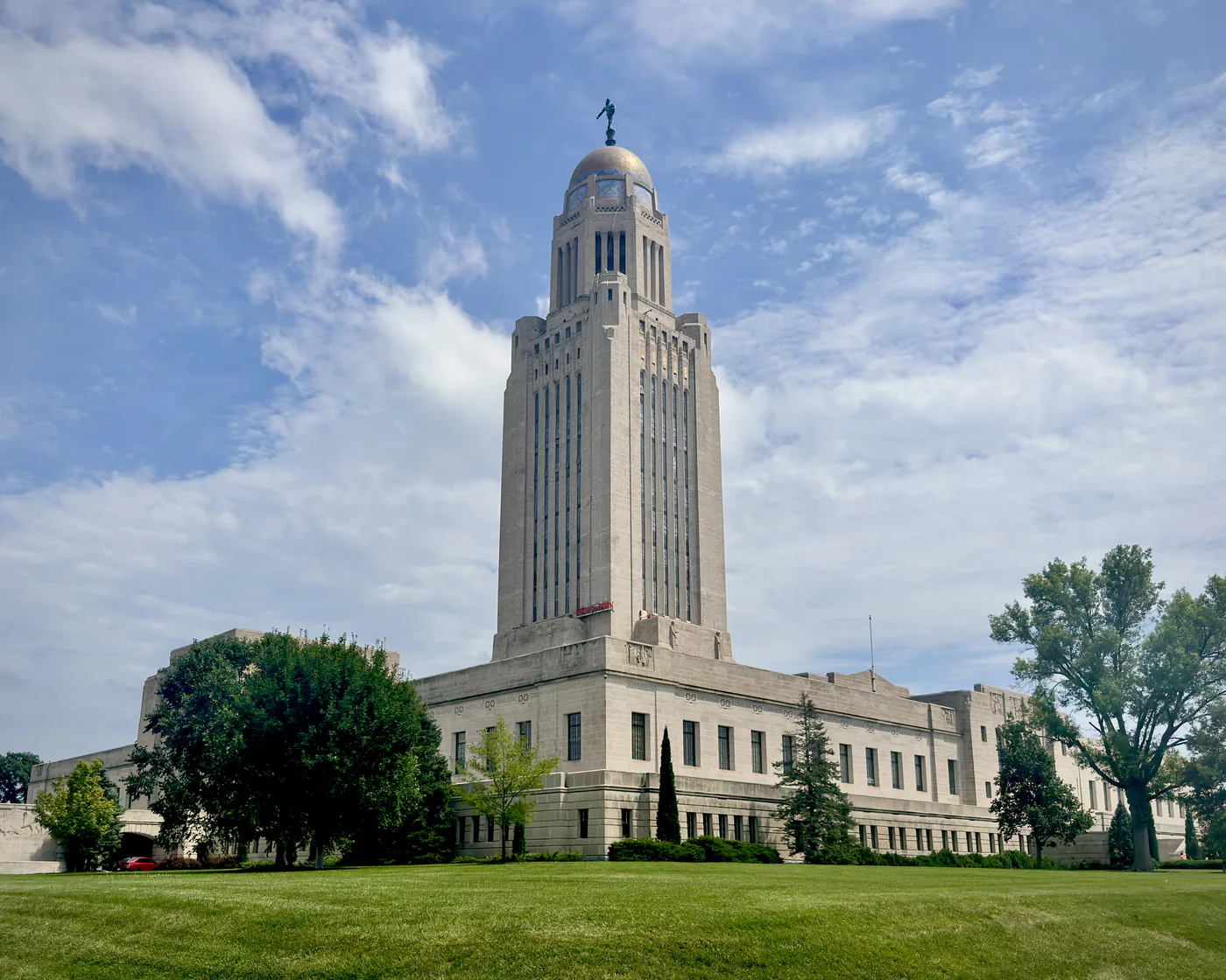The Nebraska State Capitol in Lincoln, its limestone tower and gold dome rising above a green lawn and trees under scattered cloud.