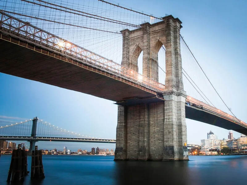 A stone tower of the Brooklyn Bridge over the East River at dusk, with the Manhattan Bridge in the distance.