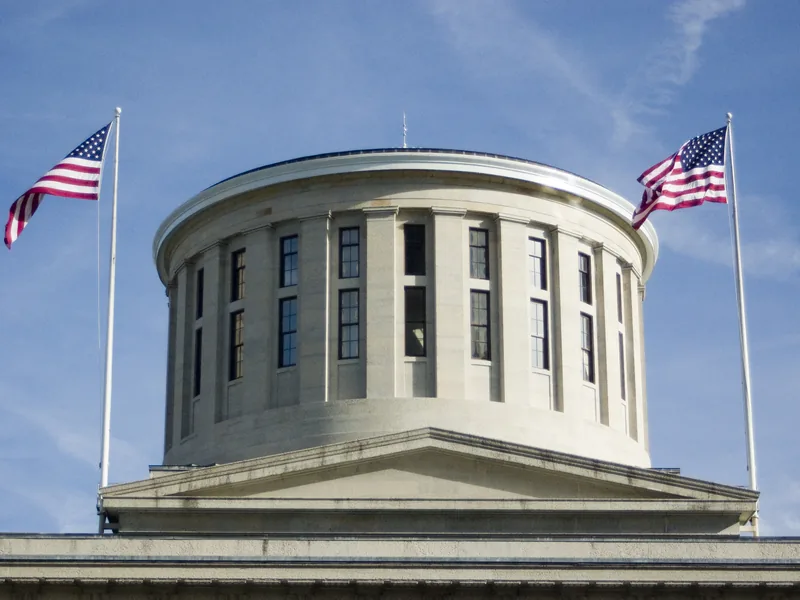 The cupola of the Ohio Statehouse in Columbus, flanked by two American flags against a blue sky.