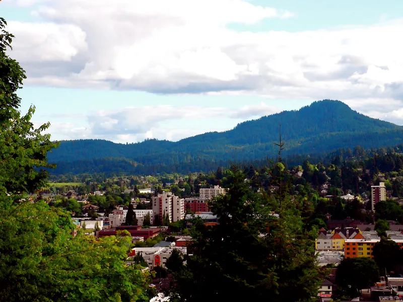 Downtown Eugene among tall evergreens, with Spencer Butte rising beyond under summer clouds.