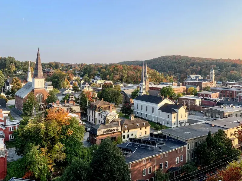 Downtown Montpelier’s church steeples and brick blocks in early autumn color, with wooded hills behind, in late-afternoon light.