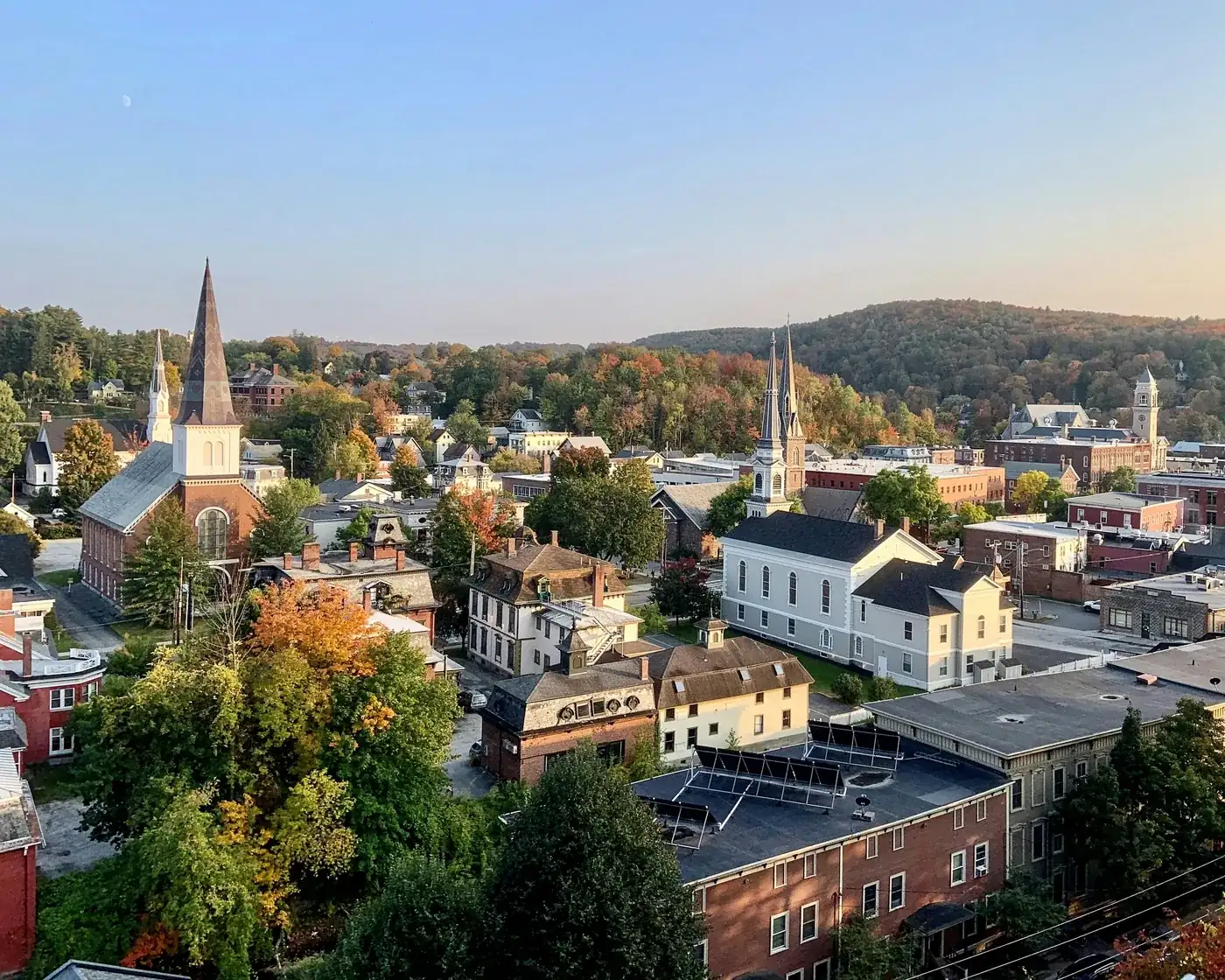 Downtown Montpelier’s church steeples and brick blocks in early autumn color, with wooded hills behind, in late-afternoon light.