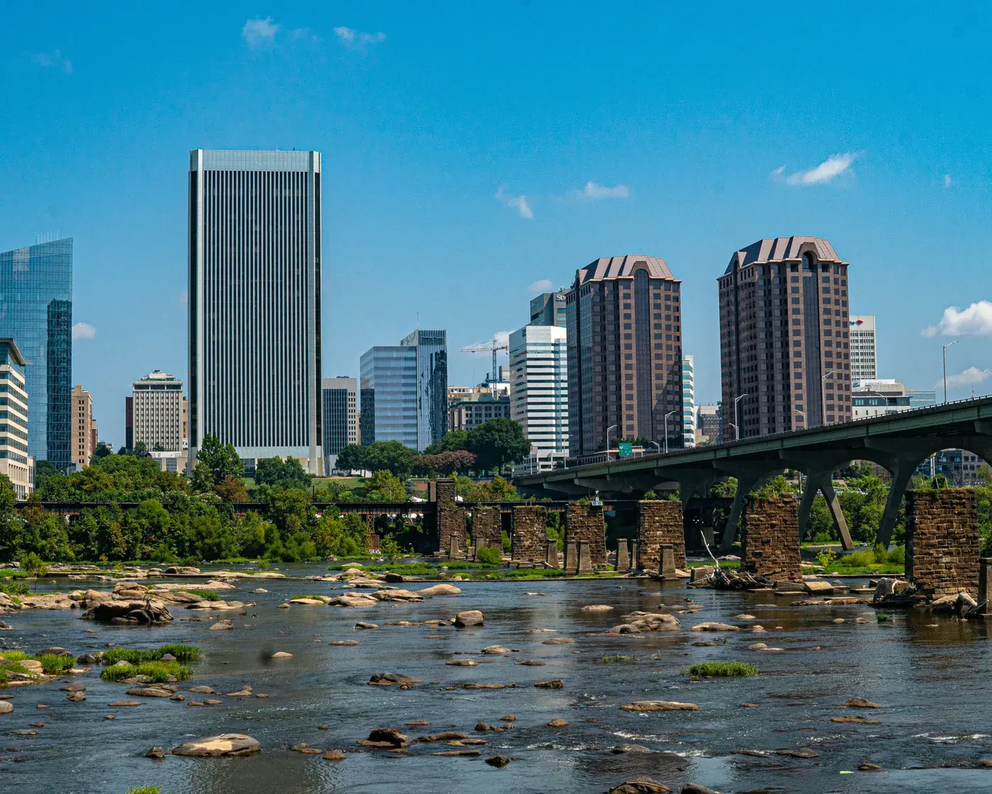 Downtown Richmond’s skyline on a clear summer day above the rocks of the James River and a row of old stone bridge piers.