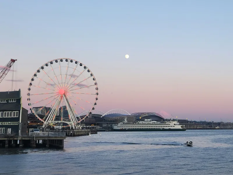 The Seattle Great Wheel on its pier at dusk, with a ferry crossing Elliott Bay and port cranes in the distance.