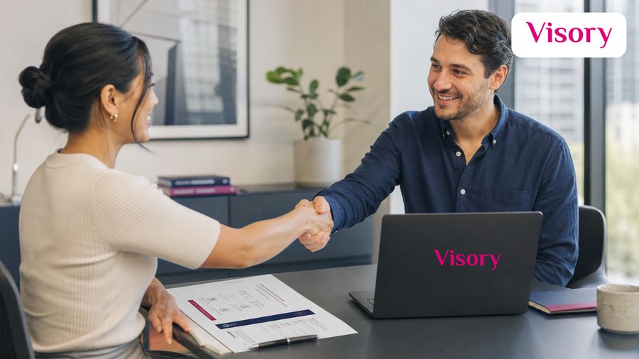 Two people shake hands across a desk beside a laptop