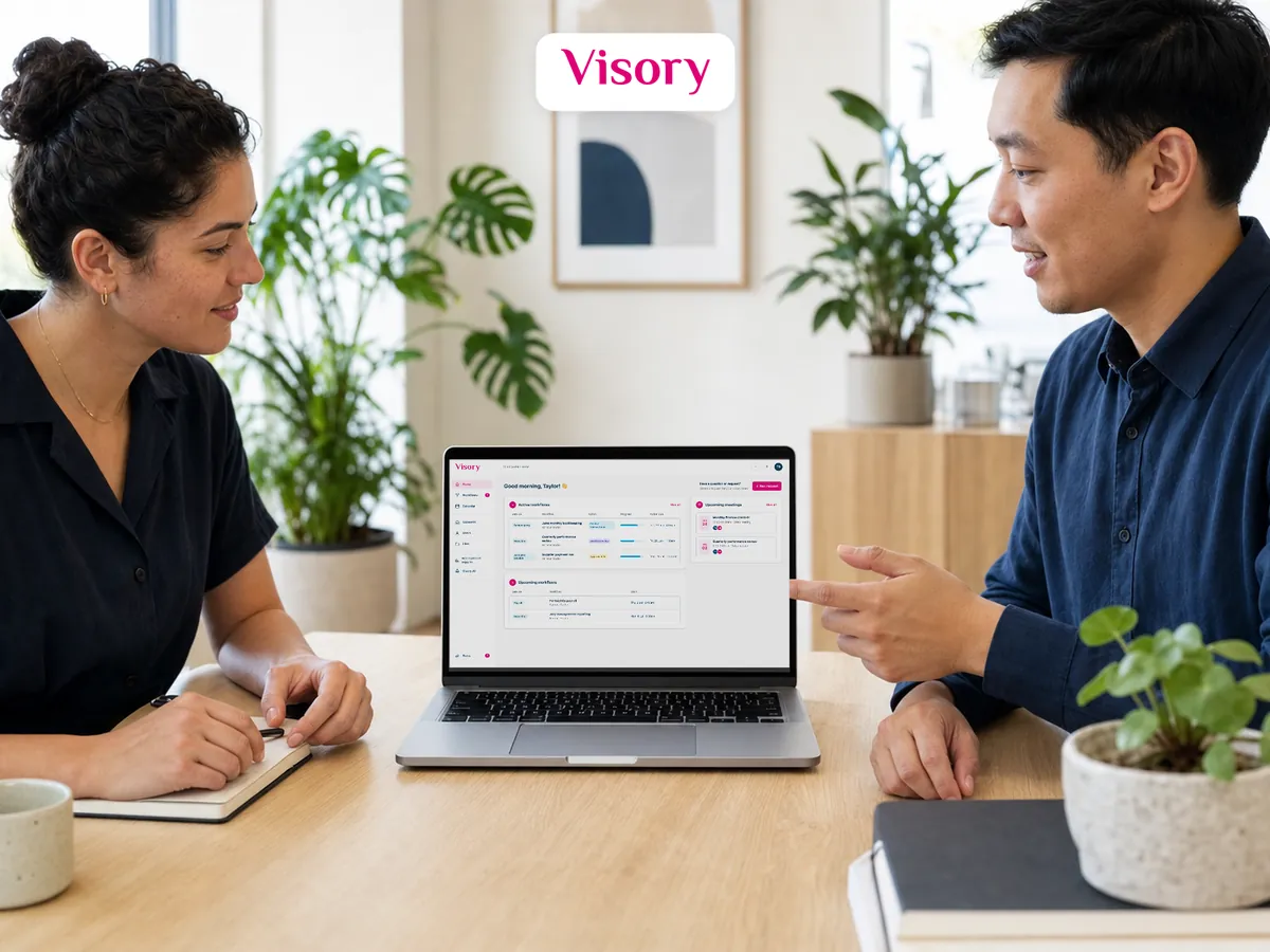 Two people at a light wood table talk over a laptop open to the Visory portal, one pointing at the screen, plants and a framed print behind them.