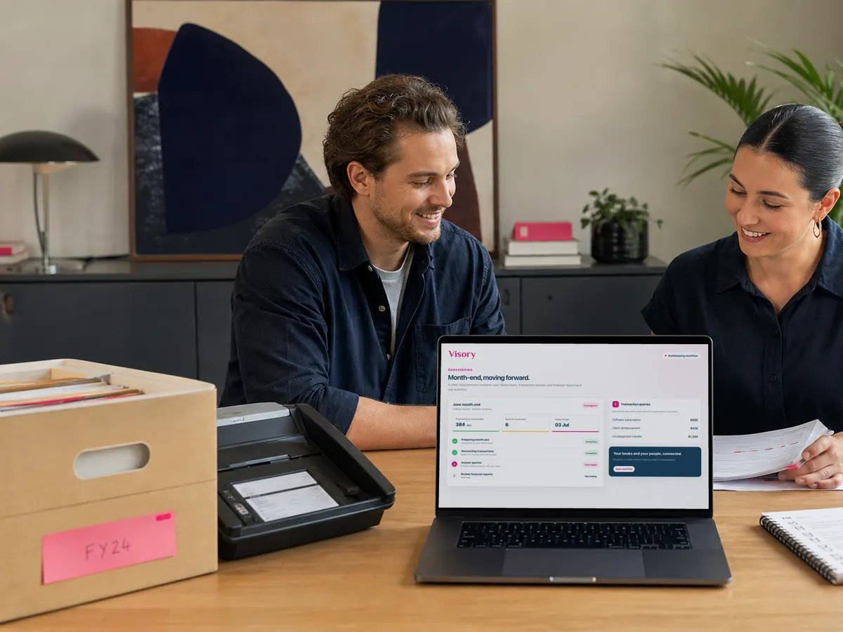 Two colleagues smile over the month-end close on a laptop, an archive box of old files and a document scanner beside them.